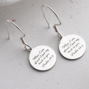 Puede incluir: Un par de pendientes plateados. Cada pendiente presenta un colgante circular con el texto grabado "When I am afraid I put my trust in you. Psalm 56:3." Los pendientes tienen un diseño de gancho.