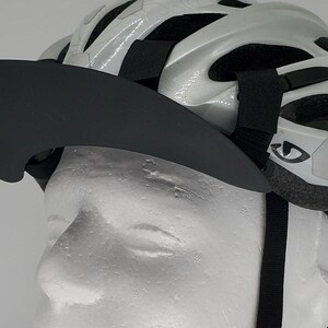 May include: A black visor with a curved brim is attached to a white bicycle helmet with black straps. The helmet has a reflective sticker with a stylized eye design.
