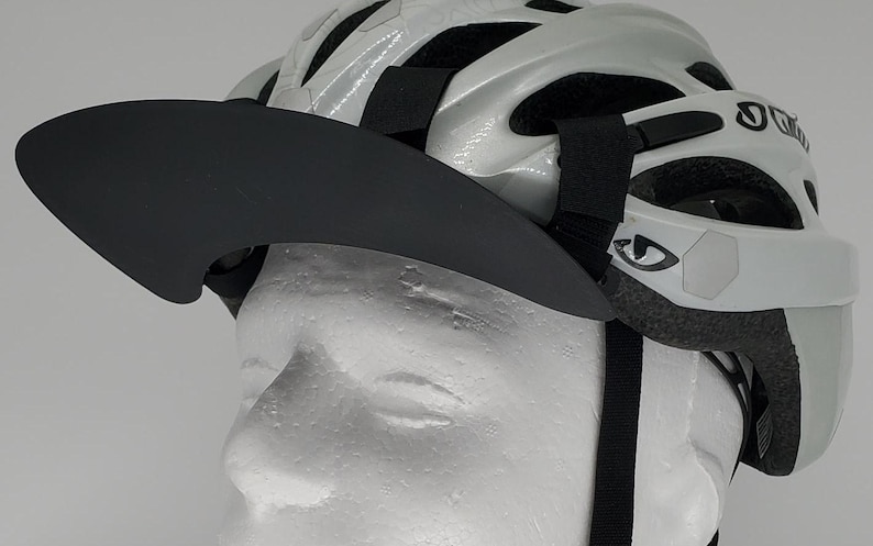 May include: A black visor with a curved brim is attached to a white bicycle helmet with black straps. The helmet has a reflective sticker with a stylized eye design.