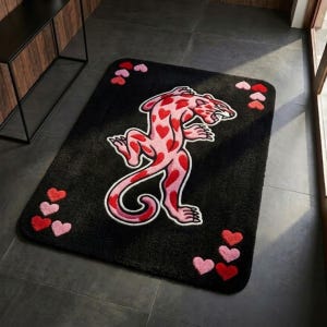 Può includere: Tappetino da bagno rettangolare nero con un disegno di leopardo rosa e macchie rosse a forma di cuore. Il tappetino ha un contorno bianco ed è circondato da piccoli cuori rosa e rossi. Un accessorio decorativo per il bagno.