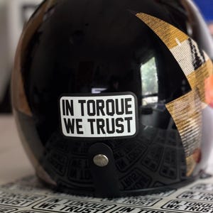Puede incluir: Un casco de motocicleta negro brillante con una pegatina blanca rectangular que contiene el texto negro IN TORQUE WE TRUST. Una correa de cuero negro con un botón de presión de metal plateado está centrada bajo la pegatina, junto a un diseño dorado.