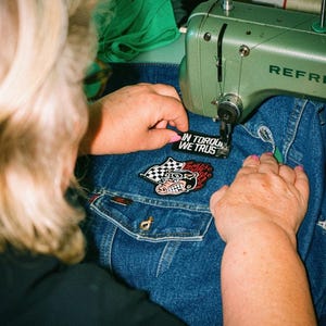 Könnte beinhalten: Eine salbeigrüne Nähmaschine näht einen schwarzen rechteckigen Aufnäher mit dem weißen Text IN TORQUE WE TRUST auf eine blaue Jeansjacke. Unter dem Aufnäher befindet sich ein buntes gesticktes Emblem mit Zielflagge und Schweinekopf.