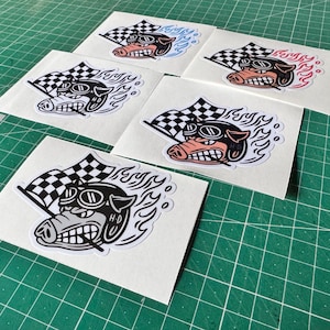 Può includere: Una collezione di adesivi con un maiale dei cartoni animati con occhiali e casco, che tiene una bandiera a scacchi. Il maiale è circondato da fiamme, con alcuni adesivi con accenti blu o rossi. Gli adesivi sono su carta bianca.