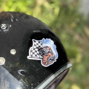 Op de afbeelding: Zwarte motorhelm met een sticker met een cartoonvarken met een bril en een geblokte vlag. Het varken heeft een vurig blauw ontwerp erachter. De helm heeft een zilveren rand en zilveren klinknagels.