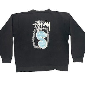 May include: Black sweatshirt with a white graphic of a blue squiggly shape inside a circle with the word "Stussy" above it.
