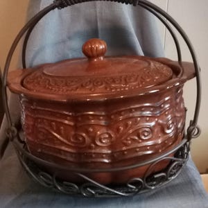 Puede incluir: Una olla de cerámica esmaltada marrón con tapa a juego, que presenta patrones en relieve de volutas y puntos. La olla se sostiene dentro de un soporte de alambre de metal oscuro que incluye un asa alta y arqueada con agarre negro texturizado.