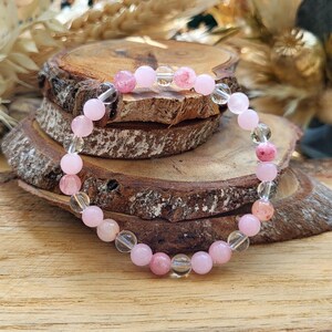 Könnte beinhalten: Ein Armband aus rosa und klaren Quarzperlen. Das Armband besteht aus runden, polierten Steinen in verschiedenen Rosa- und Klar-Tönen, die auf einem transparenten elastischen Band aufgereiht sind. Das Armband wird auf einer Holzoberfläche präsentiert.