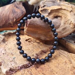 Puede incluir: Un brazalete negro de cuentas hechas de piedras redondas, lisas y pulidas. El brazalete está sobre una superficie de madera.