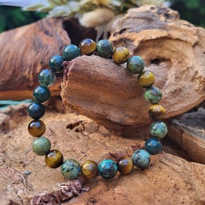 Könnte beinhalten: Ein Armband aus grünen und braunen Edelsteinperlen. Die Perlen sind rund und haben eine polierte Oberfläche. Das Armband liegt auf einer Holzoberfläche.