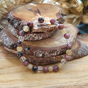 May include: A beaded bracelet featuring a variety of gemstones, including pink, yellow, and clear beads. The bracelet is displayed on a rustic wooden slice, showcasing the natural colors and textures of the stones.