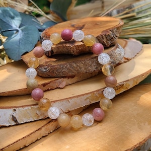 Pode incluir: Uma pulseira de contas com pedras preciosas rosa, amarelas e transparentes. As contas são enfiadas juntas. A pulseira é exibida em uma superfície de madeira, com elementos naturais ao fundo.