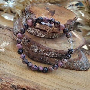 Puede incluir: Una pulsera de cuentas con cuentas de piedras preciosas rosas, negras y transparentes. La pulsera se exhibe sobre una superficie de madera rústica, mostrando los colores y texturas naturales de las piedras preciosas.