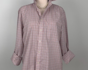 Puede incluir: Una camisa abotonada a cuadros rojos, blancos y azules con cuello con volantes y mangas largas. La camisa se exhibe en un maniquí. Las mangas están dobladas en las muñecas. Un collar de perlas está alrededor del cuello del maniquí.