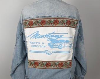 Puede incluir: Chaqueta vaquera azul claro con un panel de tapiz floral en la parte superior de la espalda. El panel presenta la palabra "Mustang" en azul claro, junto con un gráfico de coche y las palabras "Parts & Service".