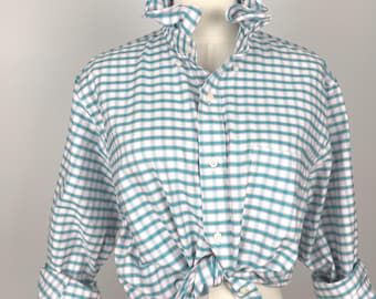 Puede incluir: Una camisa de manga larga con botones y un patrón de cuadros en color verde azulado y blanco. La camisa tiene un cuello con volantes, una tapeta de botones frontal y un bolsillo en el pecho. El dobladillo está anudado al frente y las mangas están remangadas.