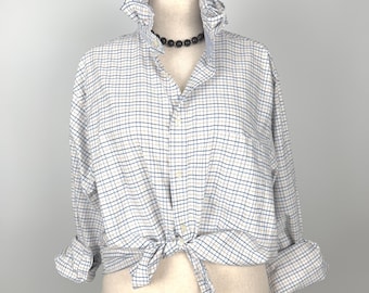 Puede incluir: Una camisa blanca con un estampado de cuadros azules y beige. La camisa tiene un cuello con volantes y está anudada a la cintura. Las mangas están enrolladas y se ve un collar de cuentas negro.