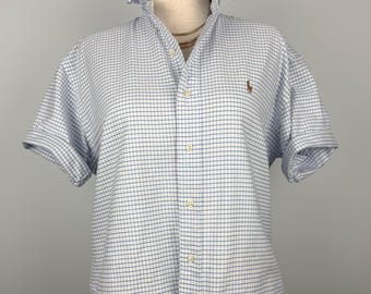 Puede incluir: Una camisa de manga corta con botones, en un patrón de cuadros azules y blancos. La camisa presenta un cuello con volantes único y mangas enrolladas. Un pequeño logotipo marrón es visible en el pecho. La camisa se exhibe en un maniquí.