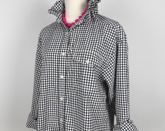 Puede incluir: Una camisa a cuadros blancos y negros con cuello y puños con volantes. La camisa de manga larga tiene un bolsillo en el pecho. Un collar de cuentas rosa brillante es visible en el escote. La camisa se exhibe en un maniquí.