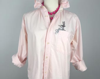 Puede incluir: Una camisa rosa claro con botones, mangas largas y un gráfico de Bugs Bunny en el bolsillo. La camisa tiene un cuello con volantes y se exhibe en un maniquí. También se ve un collar de cuentas rosas.