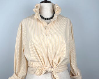 Puede incluir: Una camisa a rayas crema y blancas con cuello con volantes y mangas largas, atada a la cintura. La camisa se exhibe en un maniquí y se combina con un collar de cuentas negro.