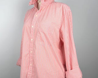 Puede incluir: Una camisa a rayas rojas y blancas con cuello con volantes y mangas largas. La camisa está abotonada por delante y tiene un bolsillo en el pecho. Las mangas están dobladas en las muñecas. La camisa se exhibe en un maniquí.