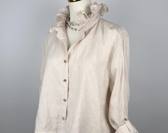 Puede incluir: Una camisa color crema con botones y rayas beige verticales. La camisa presenta un cuello con volantes y mangas largas que se pueden enrollar y asegurar con un botón. La camisa se exhibe en un maniquí.