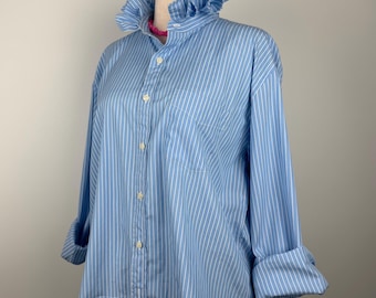 Puede incluir: Camisa a rayas azul claro y blancas con cuello con volantes y mangas enrolladas. La camisa se exhibe en un maniquí. La camisa tiene un bolsillo en el lado izquierdo.