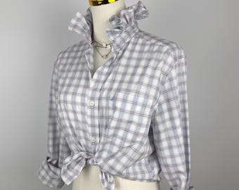 Puede incluir: Una camisa abotonada a cuadros blancos, azules y rojos con cuello con volantes y mangas largas. La camisa está anudada a la cintura y un pequeño logotipo es visible en el pecho. La camisa se exhibe en un maniquí.
