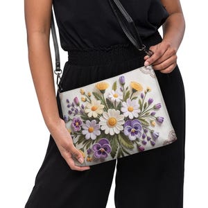 Puede incluir: Un bolso bandolera rectangular con un diseño floral. El bolso presenta un fondo blanco con un ramo de margaritas, pensamientos y otras flores en tonos amarillos, morados y blancos. El bolso tiene una correa negra y un borde negro.