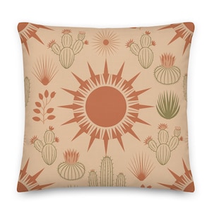 May include: Square cushion with a desert-themed pattern. The cushion features a repeating design of cacti, suns, and other desert plants in shades of orange, green, and brown on a beige background. The cushion is a square shape.