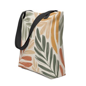 Puede incluir: Un bolso tote de lona con un estampado de hojas botánicas en tonos verdes, naranjas y beige sobre un fondo blanquecino. Tiene correas negras para llevarlo.