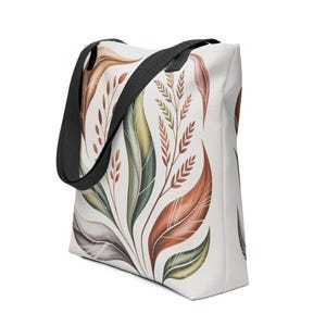 Puede incluir: Un bolso tote blanco con un diseño botánico de hojas grandes y estilizadas en tonos marrón, verde y amarillo. Tiene una correa negra y una forma rectangular.