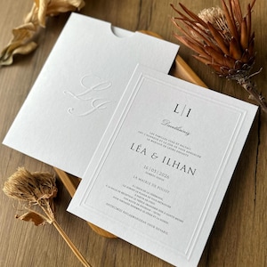 Könnte beinhalten: Elegantes weißes Hochzeitskarten-Set. Die Einladungskarte hat einen formellen schwarzen Text, einschließlich der Namen "Léa & Ilhan" und dem Datum "16/05/2026". Das Set enthält einen passenden Umschlag mit geprägten Initialen.