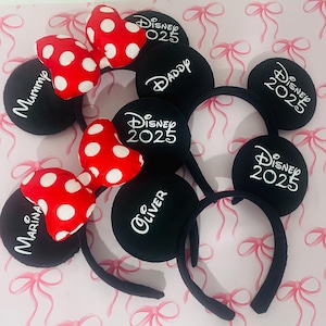 May include: Black mouse ear headbands with white text and red polka dot bows. Some headbands say "Disney 2025", "Daddy", "Mummy", "Marina", and "Oliver". The headbands are on a pink background with a bow pattern.