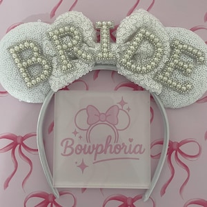 May include: A white sequined ears headband with pearl-studded letters spelling "BRIDE". The headband has a white band and a pink Bowphoria logo on a clear display. The background is pink with bow patterns.