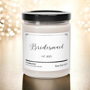 Pode incluir: Uma vela de cera de soja branca em um frasco de vidro transparente com tampa preta. O rótulo diz "Bridemaid est 2025" e "Sea Salt Orchid".