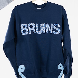 May include: A navy blue sweatshirt displaying the word "BRUINS" in white letters with a blue floral design. Light blue ribbons are tied on each side of the hem. The garment is suspended from a hanger.