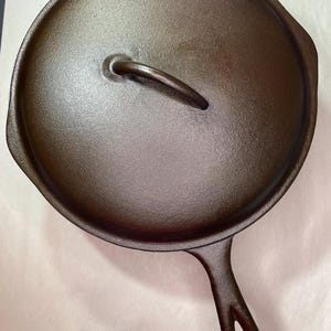 May include: A cast iron skillet with a lid. The skillet is dark brown and has a handle with a hole at the end. The lid has a curved handle. The skillet is sitting on a light-colored surface.