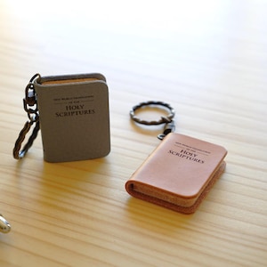 May include: Two miniature book keychains with metal rings. One is grey and the other is tan leather. Both feature the text New World Translation of the Holy Scriptures printed on the front cover. A black pen is visible on the left side of the frame.
