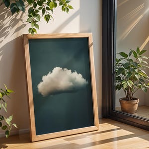 May include: A framed print of a single white cloud against a dark blue background. The frame is made of light wood and is leaning against a white wall.