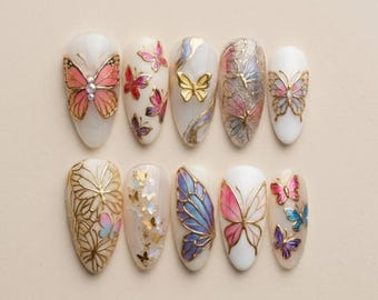 May include: A set of ten almond-shaped artificial nails, each adorned with butterfly designs. The nails feature a variety of colors, including pink, blue, gold, and purple, with intricate details and embellishments. The background is a neutral beige.