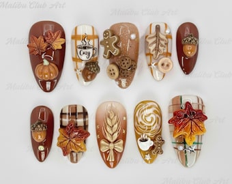 Op de afbeelding: Een verzameling van tien herfst-thema nail art ontwerpen. Elke nagel heeft een ander ontwerp, waaronder pompoenen, bladeren, eikels, peperkoekenmannetjes en koffiekopjes. Het kleurenpalet bevat tinten bruin, oranje en wit.