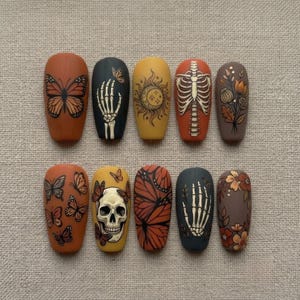 May include: A set of ten press-on nails featuring matte finishes in shades of orange, yellow, brown, and black. Designs include monarch butterflies, a human skeleton hand, a sun and moon motif, a rib cage, floral arrangements, and a human skull.
