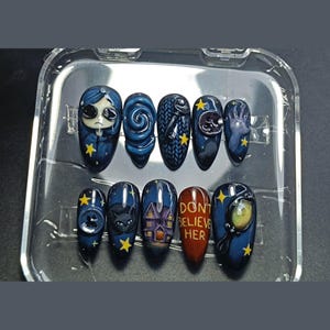 May include: A set of ten custom-designed nail art pieces. Each nail features a dark blue base with various illustrations, including a character, swirls, a cat, a house, and the text "DON'T BELIEVE HER". Accented with yellow stars.