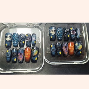 May include: A collection of press-on nails in dark blue and black. Each nail is decorated with individual artwork, including a character, swirling patterns, a house, and the text "DON'T BELIEVE HER". Yellow stars are used as accents.