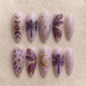 May include: A set of ten almond-shaped press-on nails in a soft lavender base color. The designs feature intricate gold and purple hand-painted details including dragonflies, crescent moons, celestial phases, and delicate floral vines with gold foil accents.