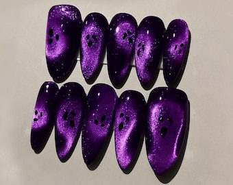 May include: A set of ten oval-shaped, purple press-on nails. Each nail features a shimmering, cat-eye effect with dark spots. The nails are arranged in two rows of five, showcasing a glossy finish and a unique design.