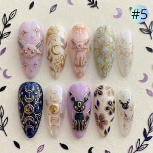 May include: A set of ten almond-shaped press-on nails featuring intricate gold-toned relief designs, including celestial motifs, floral patterns, and stylized creature illustrations in shades of lavender, white, navy blue, and mint green.
