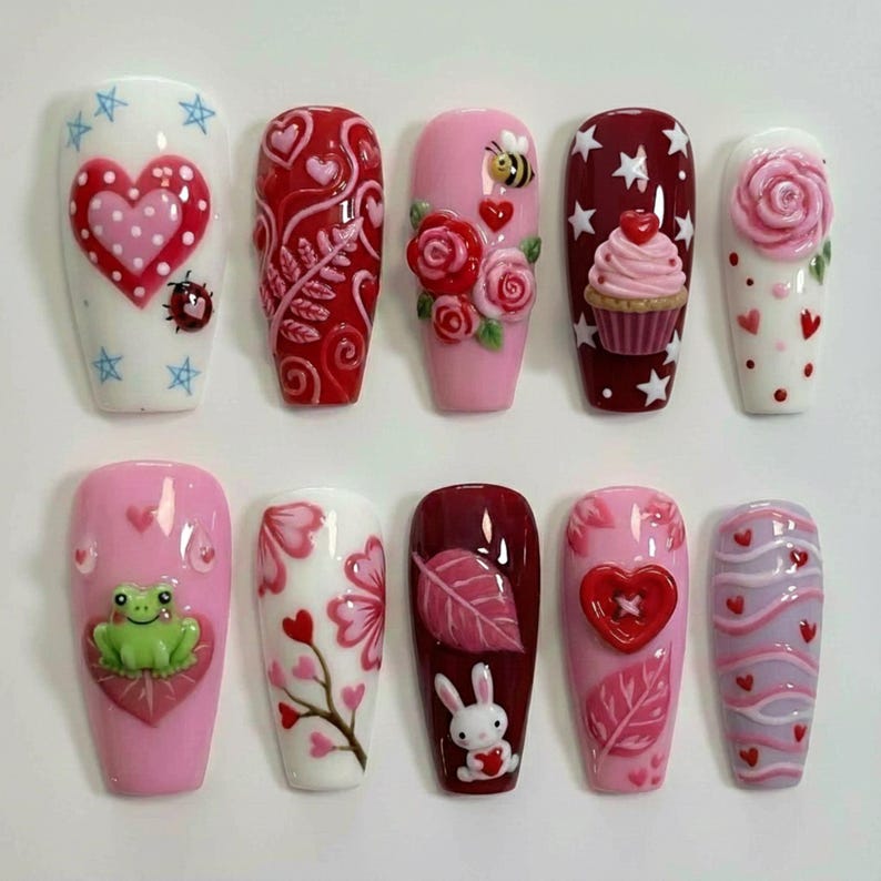 May include: A collection of ten artificial nails with various designs. The nails feature hearts, roses, a cupcake, a bee, a frog, and a bunny. Colors include red, pink, white, and burgundy. Some nails have 3D elements, while others have painted details.