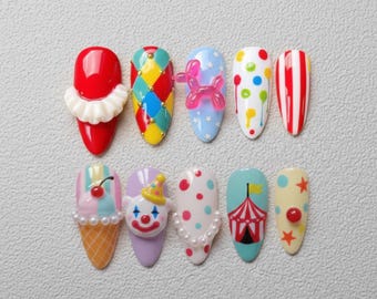 May include: A set of ten artificial nails with a circus theme. Designs include a clown, a balloon dog, a circus tent, and colorful patterns. The nails are painted in red, blue, yellow, and white.